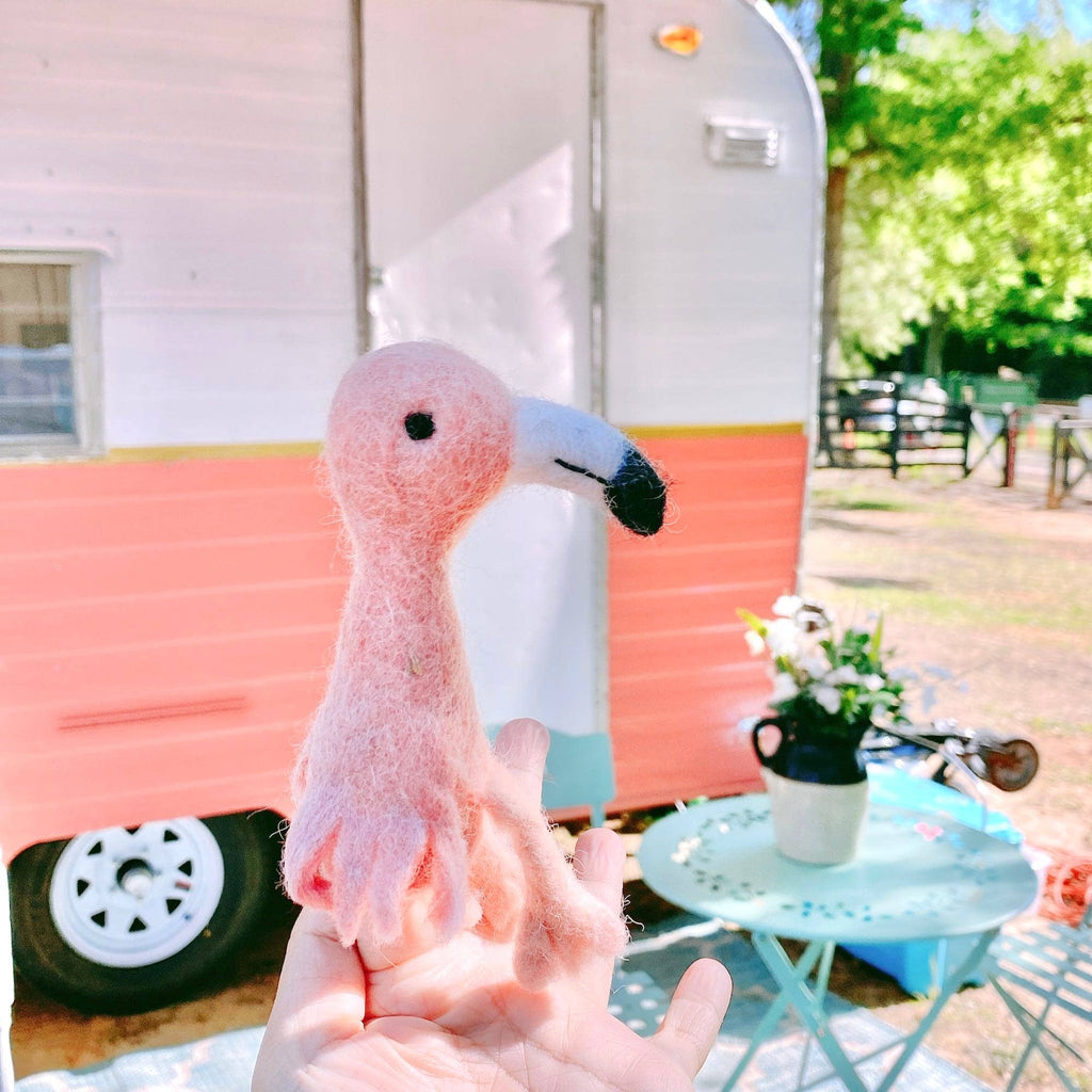 A hand displays the Flamingo Finger Puppet in front of a pastel pink and white camper. Nearby, a blue table with chairs and a potted plant sits on green grass—an ideal setting for imaginative play.