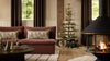 Coral & Tusk Christmas Cheer Lumbar Pillow, a rectangular embroidered holiday pillow featuring woodland animals and Christmas trees on a sofa next to a decorated tree and fireplace.