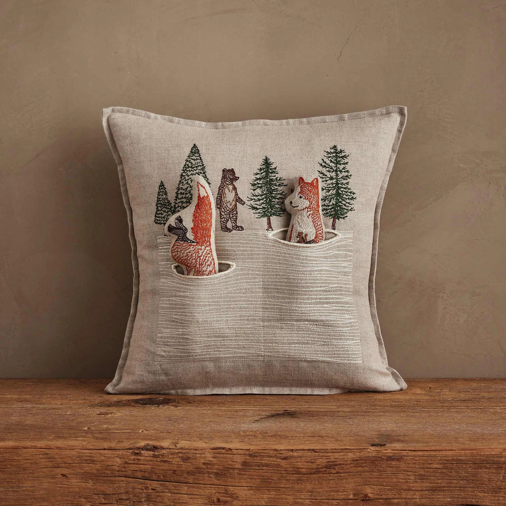 The Coral & Tusk Winter Foxes Pocket Pillow showcases embroidered fox, bear, and wolf figures among pine trees with snowy accents—ideal winter fox decor on a wooden surface.