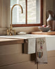 A farmhouse kitchen sink with a brass faucet and wooden countertops is decorated with the Coral & Tusk Deck the Halls Bear Tea Towel—a charming embroidered holiday accent, perfect for festive kitchen decor near the window.