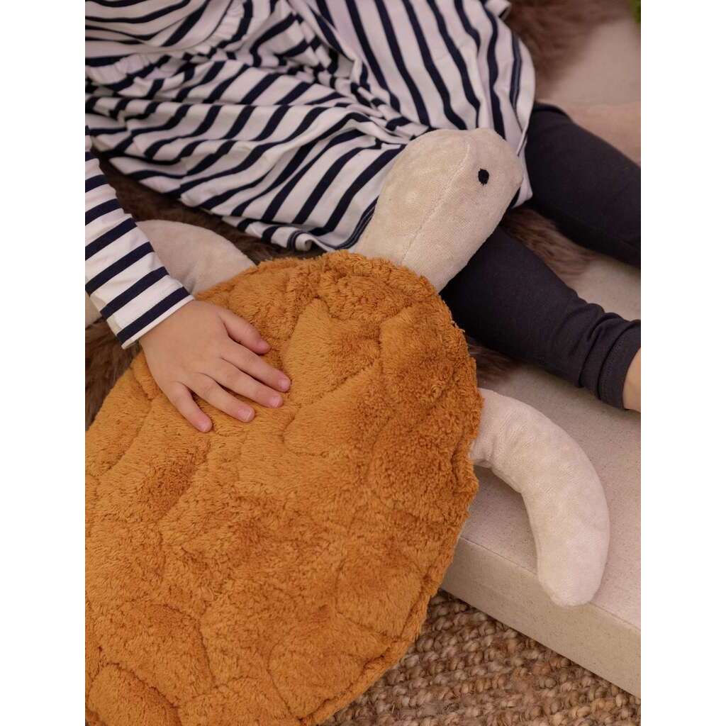 A child in a black and white striped dress sits on a mat, gently resting a hand on the Senger Naturwelt Cuddly Animal - Turtle, which is made with soft organic cotton and filled with a grain pillow.
