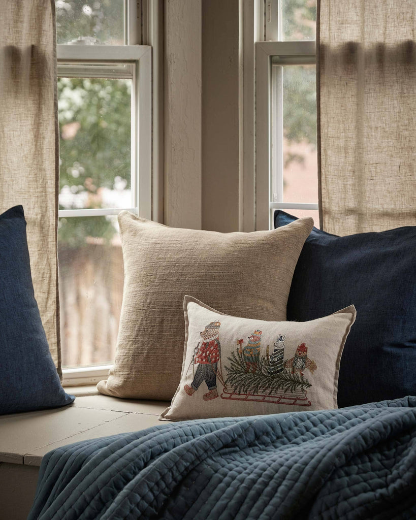 A rectangular Coral & Tusk Christmas Tree Sled Pillow made of embroidered linen, featuring a woodland scene of animals pulling a sled loaded with a Christmas tree.