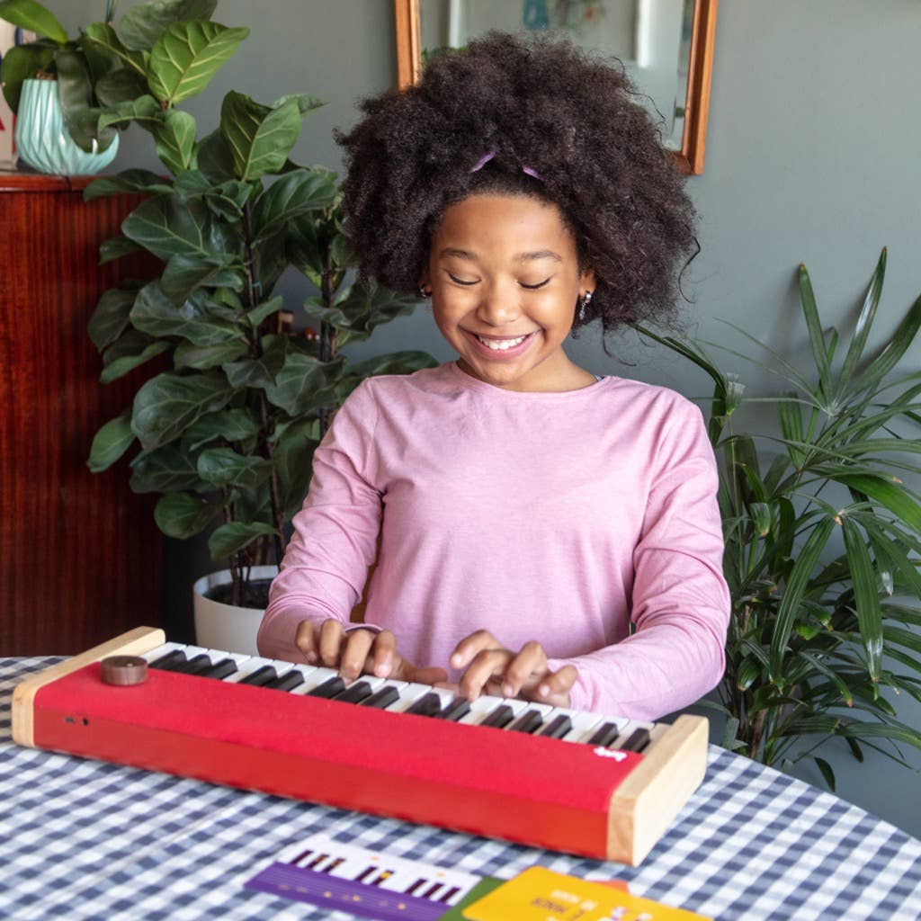 A smiling girl with curly hair in a pink shirt plays the Loog Piano, a small red portable digital piano with velocity-sensitive keys, at a checkered table surrounded by green plants and a framed picture in the background.