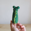 A hand with pink-painted nails holds the Felt Gator Finger Puppet, a small green wool figure with two round ears and a wavy yellow line down its back, ideal for imaginative play against a softly lit plain background.