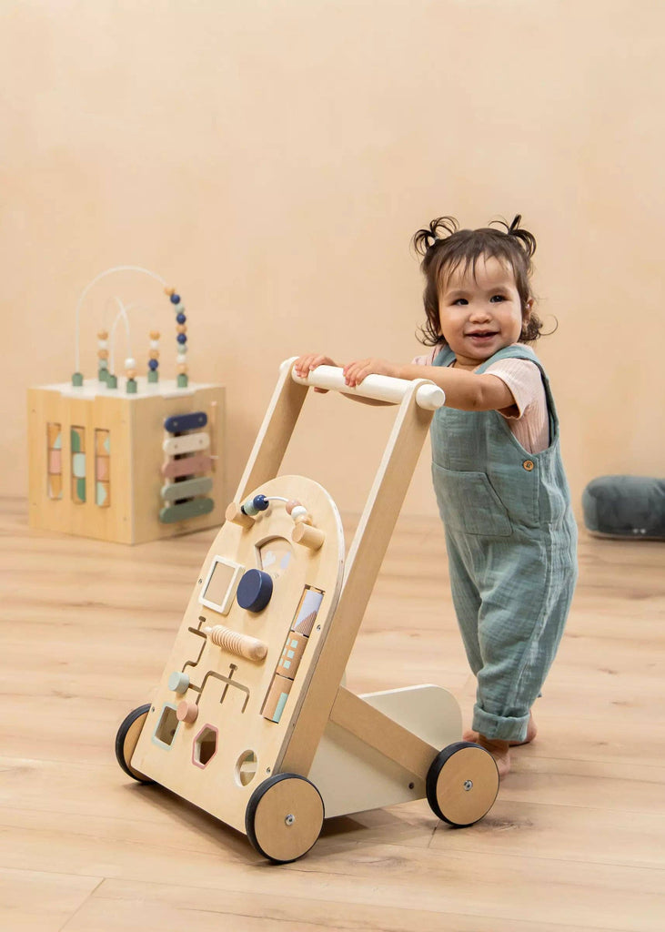 A smiling toddler in blue overalls stands on a wooden floor, holding the Wooden Activity Walker. A baby walker and a wooden activity cube toy, designed as engaging sensory toys, are visible in the background.
