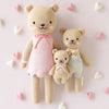 The Cuddle + Kind Baby Honey Bear (Lavender Bow), a hand-knit, heirloom-quality doll, is displayed on a light background with pastel heart decorations for a charming nursery accent.