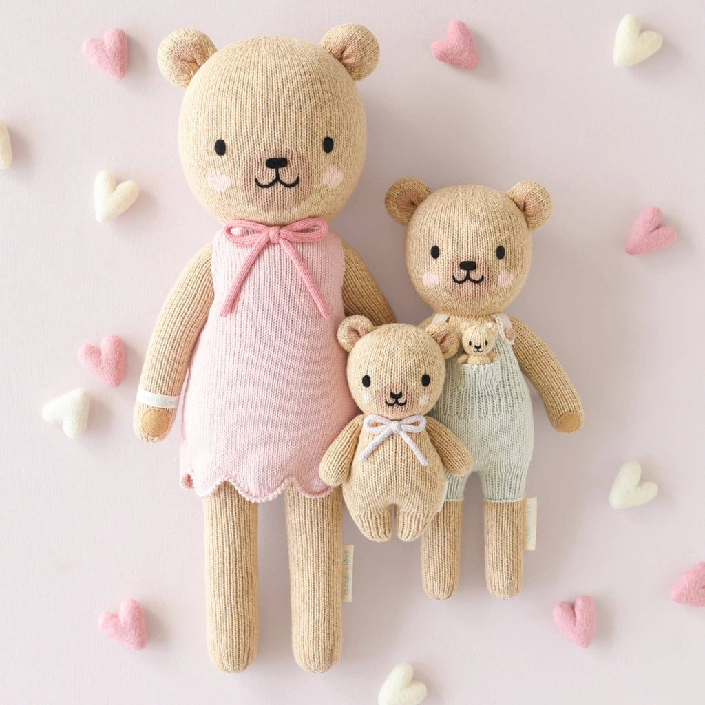 The Cuddle + Kind Baby Honey Bear (Lavender Bow), a hand-knit, heirloom-quality doll, is displayed on a light background with pastel heart decorations for a charming nursery accent.