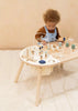 A toddler with curly hair plays intently with a bead maze and various wooden toys on a Wooden Activity Table in a softly lit room with a beige background.