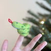 The Snake Finger Puppet features green fabric with black spots and a red tongue, wrapping around your finger for imaginative play. A blurred green Christmas tree in the background inspires creative storytelling moments.