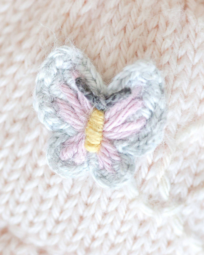 A close-up of a small, embroidered butterfly in purple, yellow, and white thread on Cuddle + Kind Kelsie the Pony—a soft, pale pink hand-knit doll, handmade in Peru under fair trade standards.