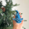 A hand holds a Finger Puppet: Whale Shark, a small blue puppet with white spots, in front of a blurred green pine tree, bringing marine magic to life.