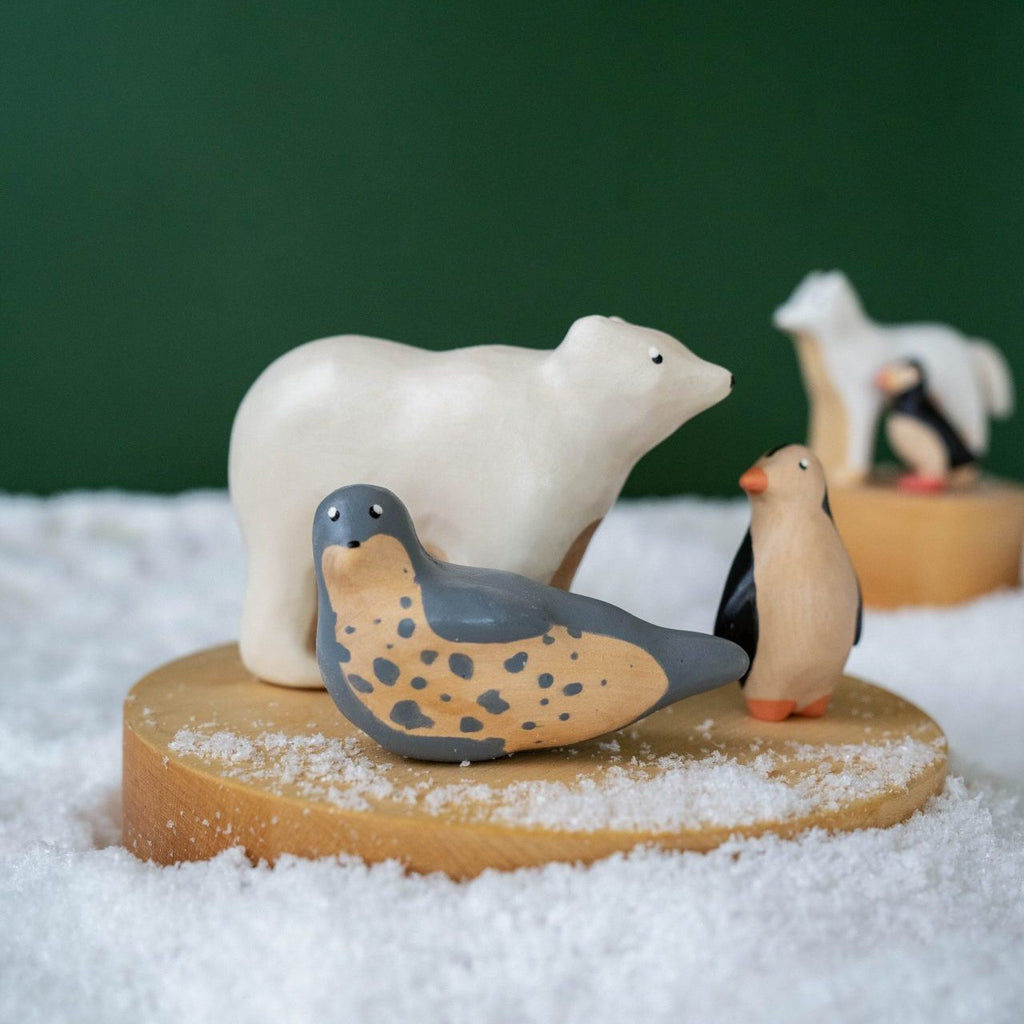 The Handmade Wooden Polar Bear, along with a penguin and spotted bird, is artistically displayed on a round wooden platform. Made from natural wood with child-safe paint, another similar platform features these figures against a green backdrop resembling snow.
