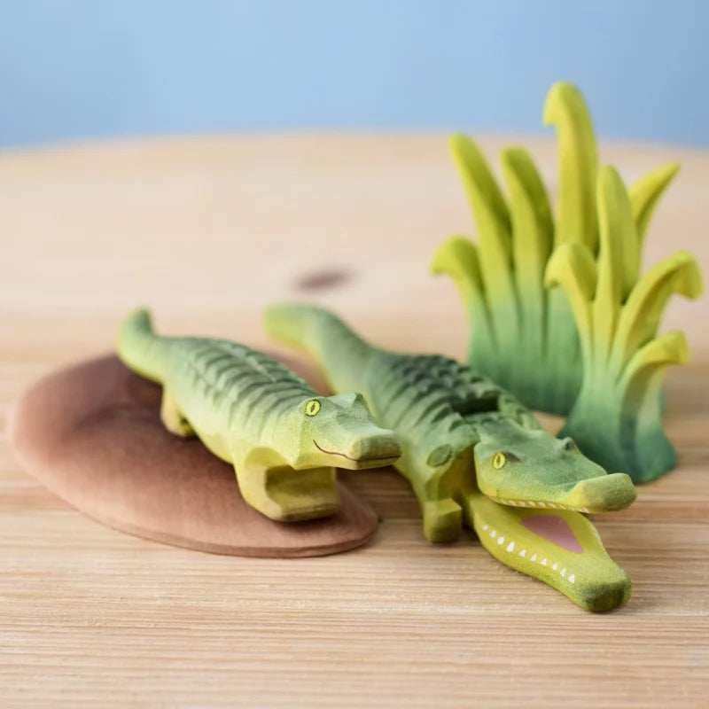Two wooden crocodiles and green reeds from the Bumbu Wooden Crocodile Cove Set, crafted from sustainable wood, are arranged by a brown oval "pond"—perfect for Montessori and Waldorf-style play.