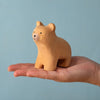 A hand holds the Bumbu Handmade Wooden Curious Bear, a small light brown toddler wooden figure, against a plain light blue background.