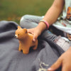 A child holds the Bumbu Handmade Wooden Curious Bear while sitting on a gray blanket outdoors, wearing a beaded bracelet. Grass in the background highlights sensory development through tactile play.
