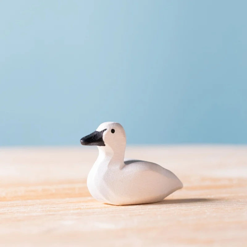The Bumbu Handmade Wooden Baby Swan, featuring a black beak, is displayed on a light wooden surface with a pale blue background.