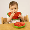 A baby in a wooden high chair eats watermelon from the Wally the Watermelon Plate & Spoon Set, with matching green dinnerware on the table in front of them.