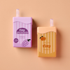 Two small juice boxes with straws from the Tiny Tummies Set - Juice Box Duo rest on a peach background—one in purple labeled “tiny tummies grape juice,” the other in orange featuring an illustrated orange and the phrase “it’s the tiny things.” Perfect as playful doll accessories, these interactive juice boxes add a charming touch to any toy collection.