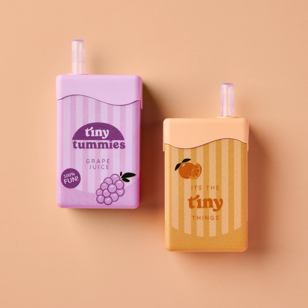 Two small juice boxes with straws from the Tiny Tummies Set - Juice Box Duo rest on a peach background—one in purple labeled “tiny tummies grape juice,” the other in orange featuring an illustrated orange and the phrase “it’s the tiny things.” Perfect as playful doll accessories, these interactive juice boxes add a charming touch to any toy collection.