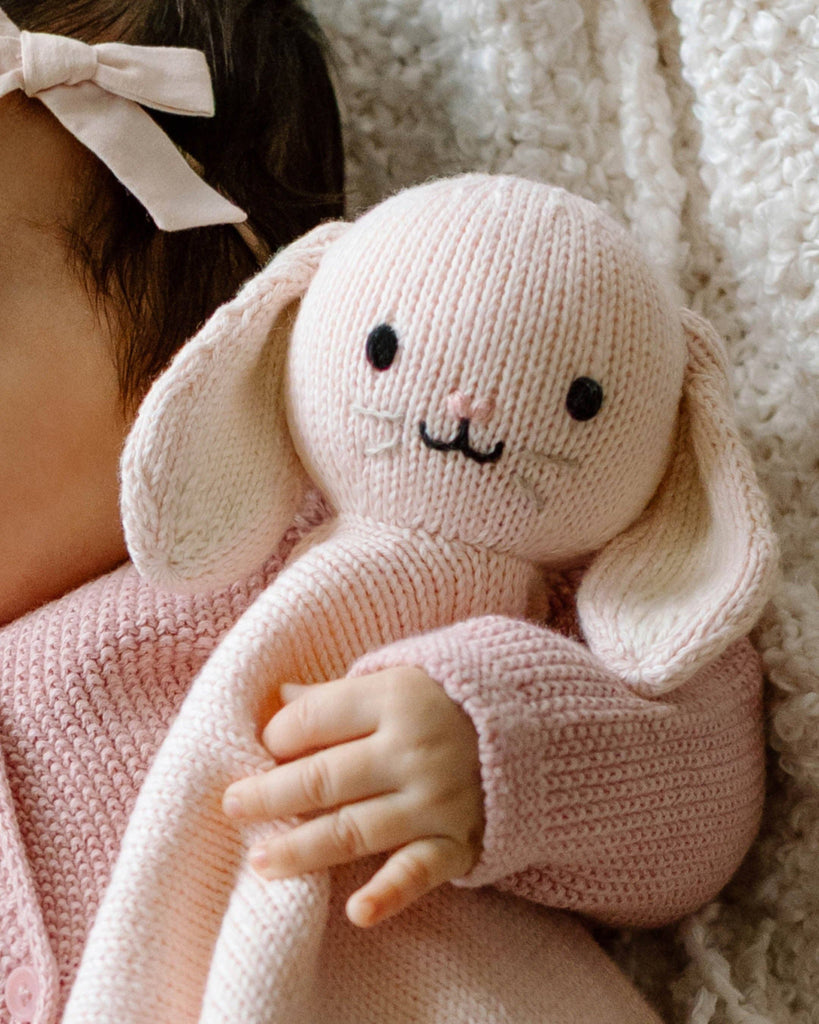 A baby in a pink sweater and bow holds the Cuddle + Kind Organic Cotton Baby Bunny Lovey (Blush)—a hand-knit pink bunny with floppy ears—while resting on a soft, textured white blanket.