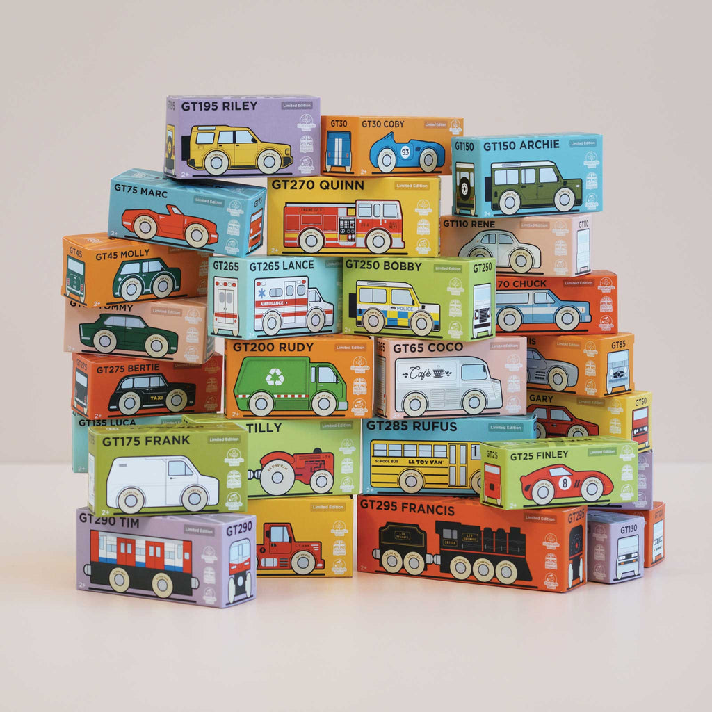 A pyramid stack of colorful toy vehicle boxes, with each box featuring the product name “No. 49 GT295 Francis | Made in England” alongside a cartoon illustration of different vehicles like buses, trucks, classic locomotives, and cars, arranged on a plain surface.