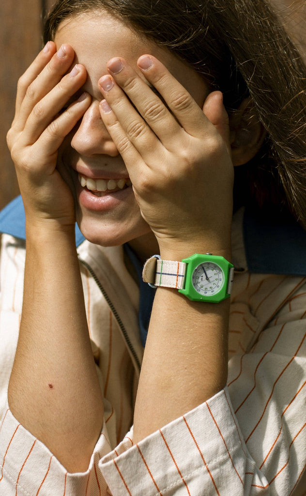 A smiling person covers their eyes with both hands, wearing a green Mini Kyomo Watch x Taxi Brousse - Color Notebook with a striped band. They have long hair and are dressed in a cream jacket with red stripes.