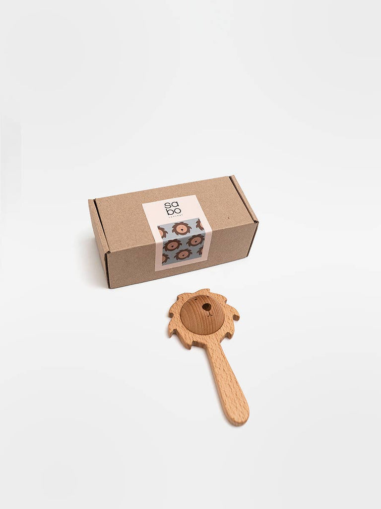 A brown cardboard box with a white label sits next to the Sabo Concept Lion Rattle - Natural, a baby toy crafted from natural materials, displayed on a white background.