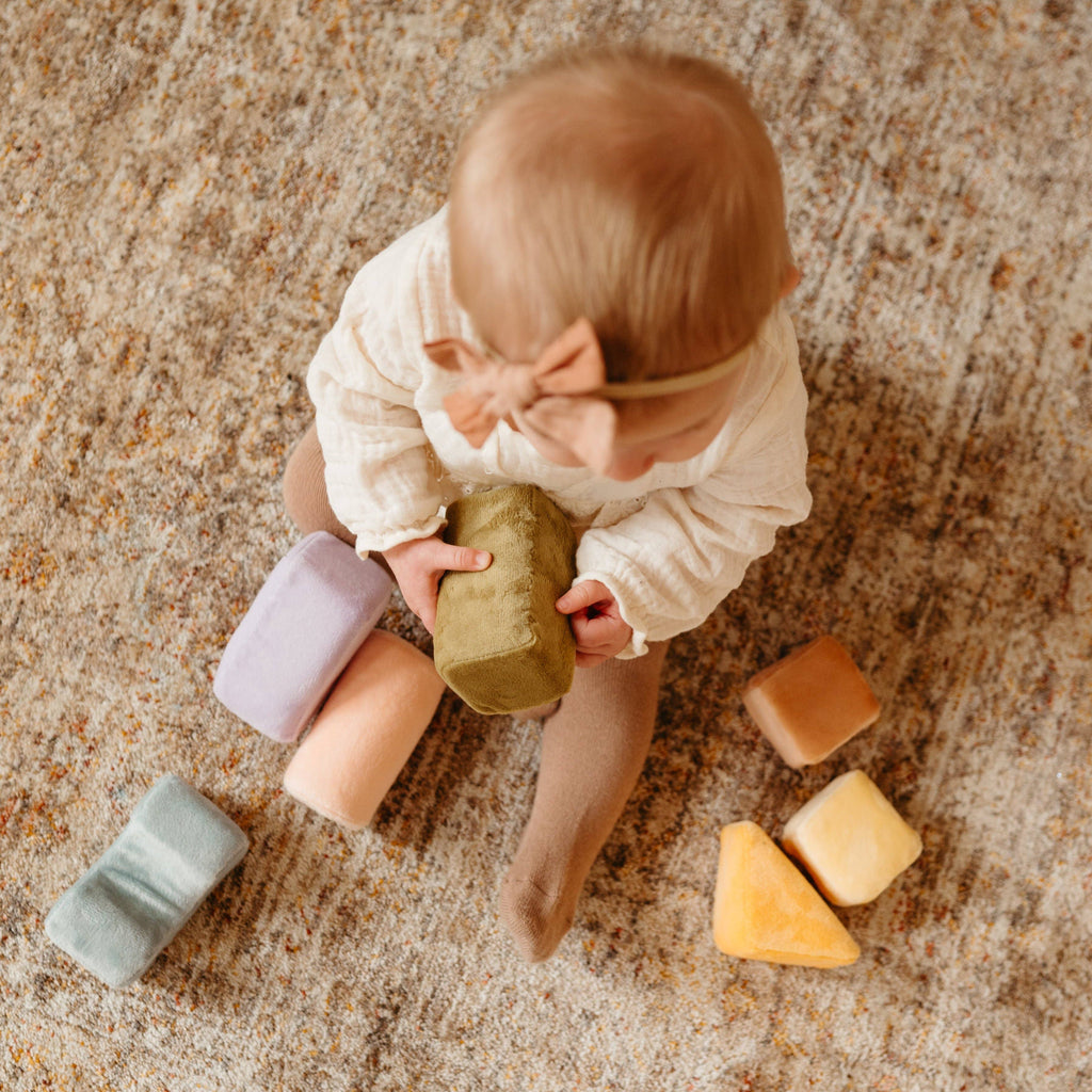 A baby wearing a headband sits on a carpet, holding a soft green Soft Toy Block and surrounded by pastel-colored Soft Toy Blocks in various shapes.