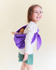 A smiling young girl in a white shirt and green shorts carries a doll in a Sarah's Silks Solid Color Playsilk - Purple sling over her shoulder, inspiring open-ended play against a light background.