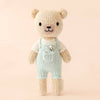 Cuddle + Kind Tiny Charlie the Honey Bear is a hand-knit doll from Peru, featuring light blue overalls, a white shirt, black embroidered features, and a gentle expression. It stands upright against a pale beige background.