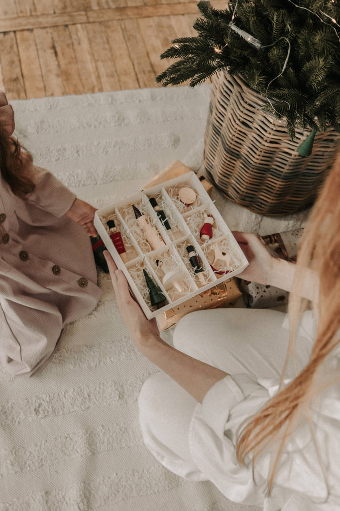 Two people sit on a light rug by a basket with a small Christmas tree. One holds an open box of Sabo Concept Christmas Ornaments while the other, dressed in pink, admires the hand-painted festive collection.