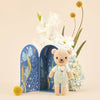 Cuddle + Kind Tiny Charlie the Honey Bear, hand-knit in pale overalls, stands before a blue floral-patterned box surrounded by white, yellow, and light blue flowers against a soft beige background.