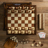 The Heirloom Edition: 7-in-1 chessboard is displayed with pieces ready for play, surrounded by dominoes, playing cards, a cribbage board, and a drawstring pouch with game pieces on a wooden surface.