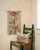 A Coral & Tusk Holiday House Advent Calendar shaped like a brick house with pockets, featuring small gifts and candy canes, next to a chair with an embroidered pillow depicting a horse-drawn sleigh.