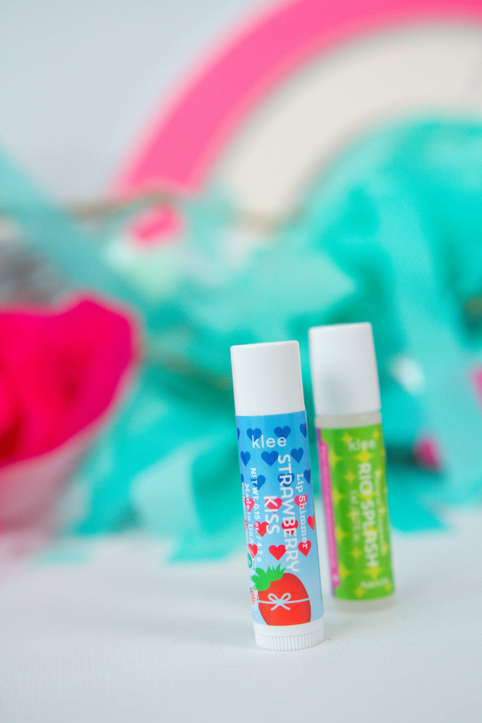 Two tubes of Klee Naturals Makeup Kit - After the Rain- Rainbow Dream labeled “Strawberry Kiss” and “Rio Splash” standing upright against a blurred colorful background.