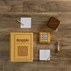 The Board Game: Boggle Vintage Bookshelf Edition is set on wood, showing its box, game cube with letter dice, timer, brown cover, two pencils, and two notepads with handwritten word lists.