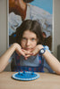 A girl with long brown hair sits at a table, wearing a blue dress, blue nail polish, and the Mini Kyomo Watch - Blue Sunday - Blue Checkered. A painting hangs on the wall behind her.