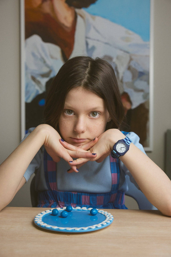 A girl with long brown hair sits at a table, wearing a blue dress, blue nail polish, and the Mini Kyomo Watch - Blue Sunday - Blue Checkered. A painting hangs on the wall behind her.