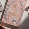 A pink hardcover "Alice in Wonderland | Collector's Edition" lies on a sunlit surface, decorated with flowers, playing cards, and illustrations of Alice and the White Rabbit.