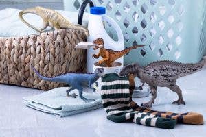 The Schleich Velociraptor is shown among laundry items, including a plastic basket, detergent, folded towels, and striped socks on the floor. In the background is a plastic laundry basket. This detailed dinosaur toy features movable arms for added play value.