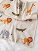 The Organic Cotton Knit Baby Blanket - Jungle, in beige with lions, elephants, giraffes, and crocodiles, rests on a white furry surface. A lion-shaped rattle sits atop this 100% organic cotton blanket for a playful touch.
