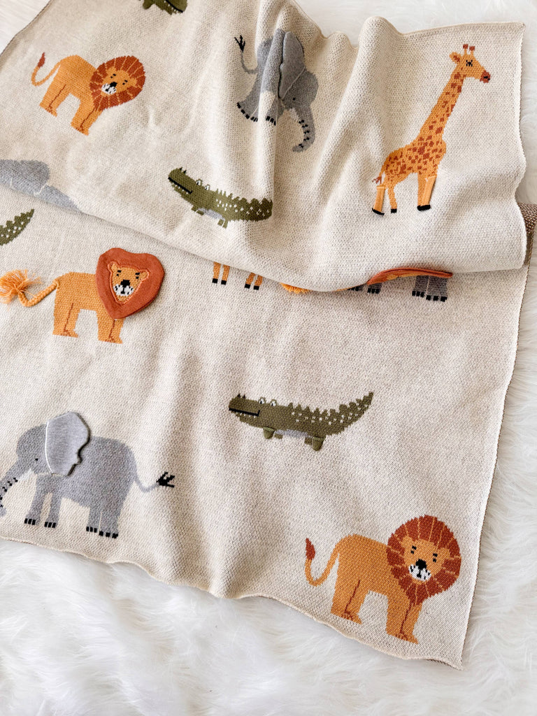 The Organic Cotton Knit Baby Blanket - Jungle, in beige with lions, elephants, giraffes, and crocodiles, rests on a white furry surface. A lion-shaped rattle sits atop this 100% organic cotton blanket for a playful touch.