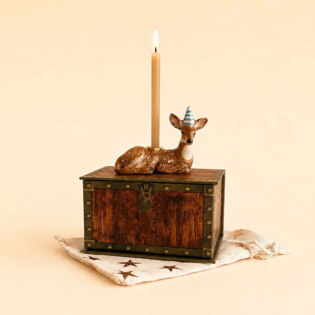 A lit candle sits on a decorative box with gold accents, while the Deer Cake Topper - Final Sale (ships in one week), wearing a blue party hat, stands nearby. The box rests on a star-patterned cloth.