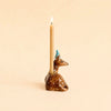 A lit candle sits in the Deer Cake Topper - Final Sale (ships in one week), a brown deer with a blue striped party hat and gold accents, against a beige background.