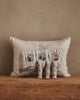 The Coral & Tusk Aspen Log Bunnies Pocket Pillow, a beige linen pillow embroidered with a log and houses, sits against a tan wall. In front, three detailed removable rabbit figurines stand on a wooden surface.