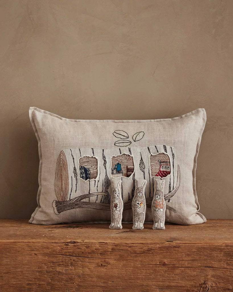 The Coral & Tusk Aspen Log Bunnies Pocket Pillow, a beige linen pillow embroidered with a log and houses, sits against a tan wall. In front, three detailed removable rabbit figurines stand on a wooden surface.
