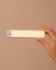 A hand holds a rectangular, battery-powered LED light with a frosted cover and a small round sensor on one end, against a plain beige background—similar to the modern lighting featured in the Fully Furnished Dollhouse With Lights, House of New York - The Six-Storey Giant.