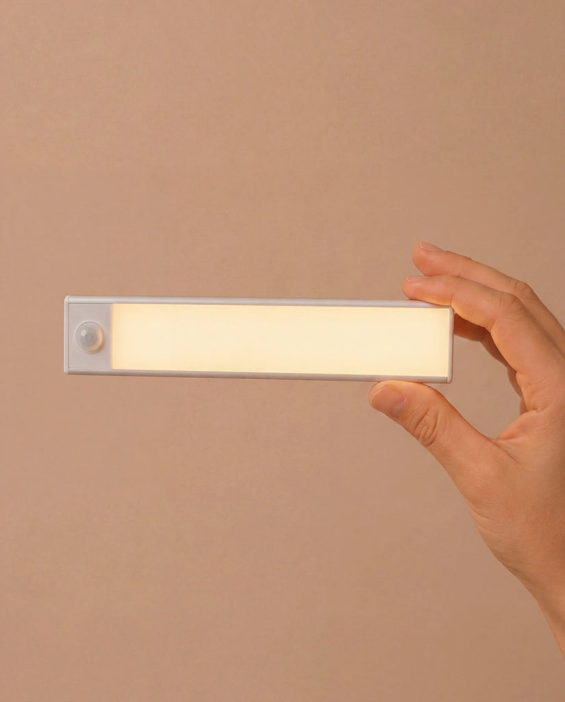 A hand holds a rectangular, slim, white LED light with a small circular sensor on one end, set against a plain beige background—an ideal accessory for illuminating every room in your House of Paris Luxury Dollhouse – A Tale of Parisian Magic, or for bringing a touch of modern elegance to this enchanting Parisian miniature home.