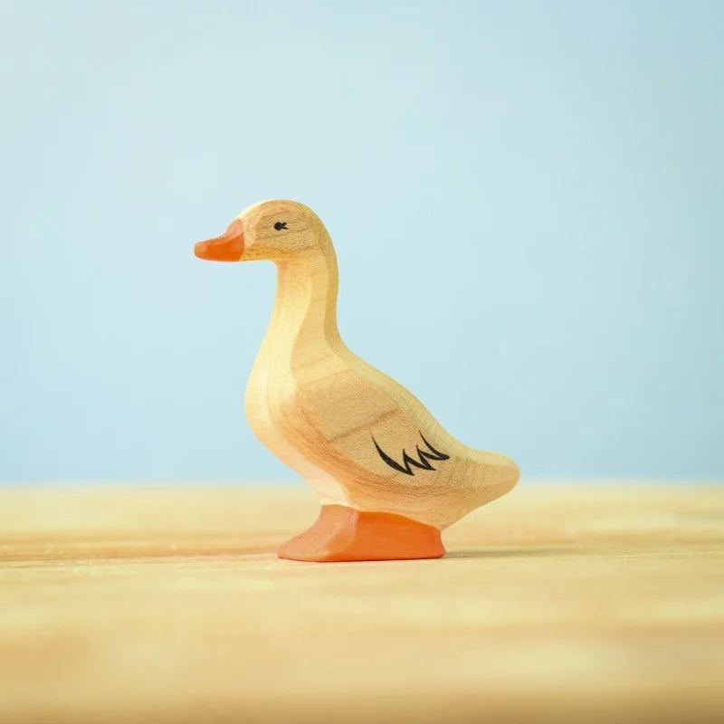 The Bumbu Handmade Wooden Domestic Duck features a small, handcrafted design with an orange beak and feet, black detailing on the eyes and wing, standing on a light wooden surface against a pale blue background.