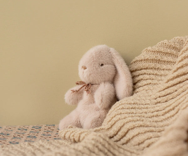 The Maileg Plush Bunny, Mini - Light Powder, a soft stuffed animal with floppy ears and a brown ribbon, sits on a textured beige knit blanket atop a patterned surface. It’s crafted from recycled polyester with a light tan wall as the backdrop.